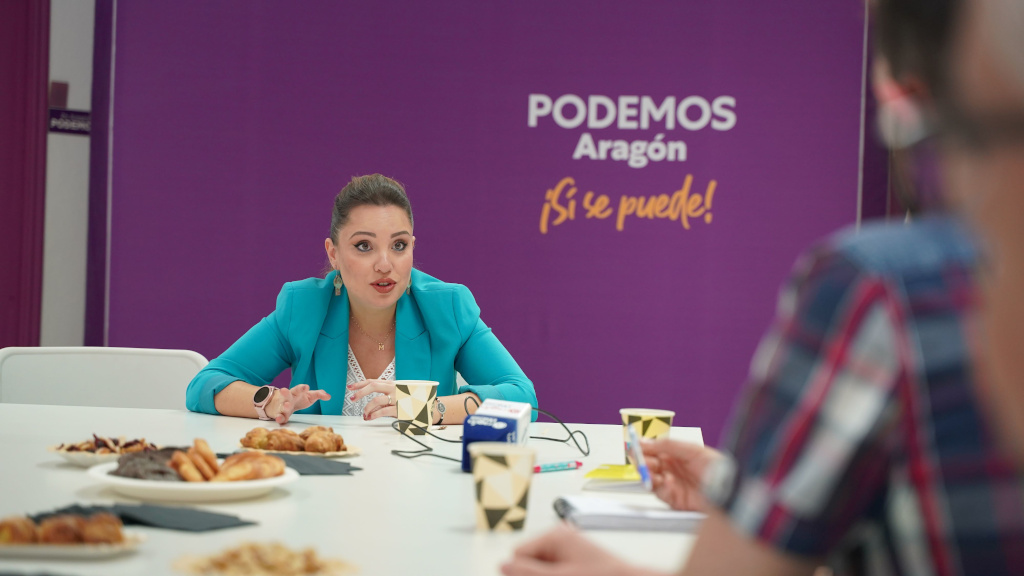 Maru Díaz de Podemos Aragón, sentada en una mesa con el logo de podemos al fondo, con un desayuno al que ha invitado a los periodistas