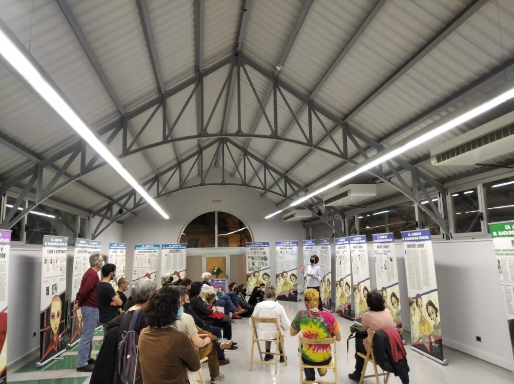 La exposición sobre la mujer en el anarquismo en la sede de CGT con varios paneles y varias personas sentadas durante la presentación