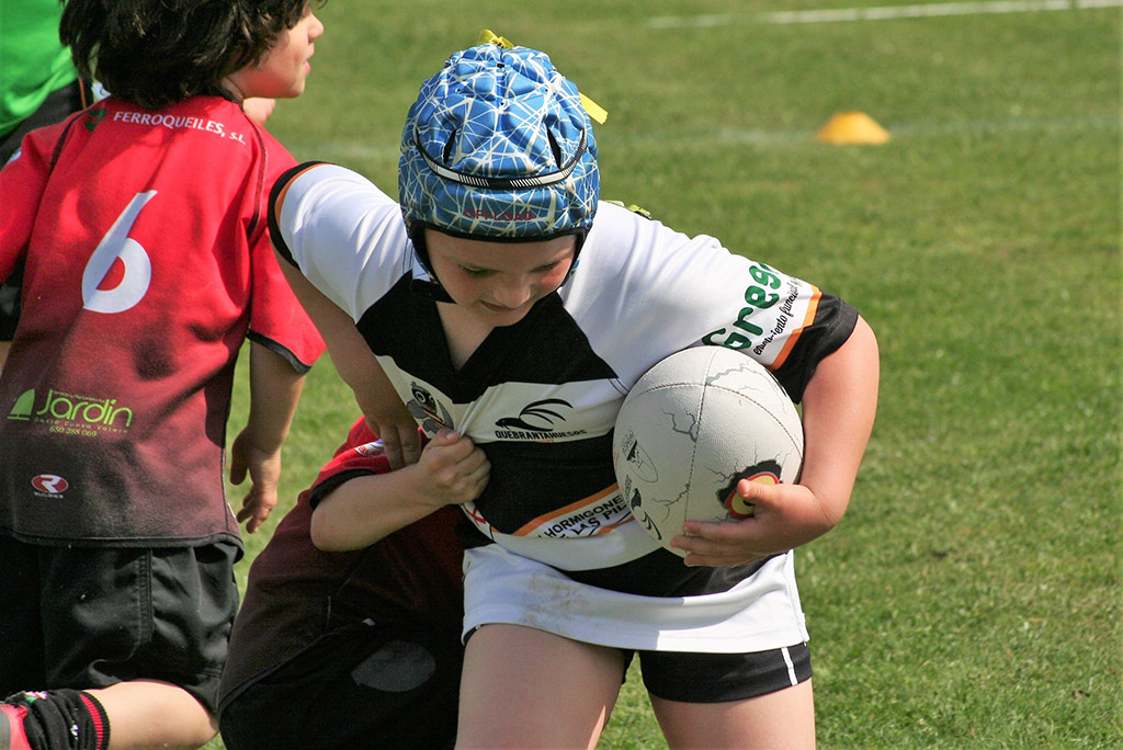 Un joven ataviado con uniforme de rugby juega un partido con otros chavales en un campo con cesped