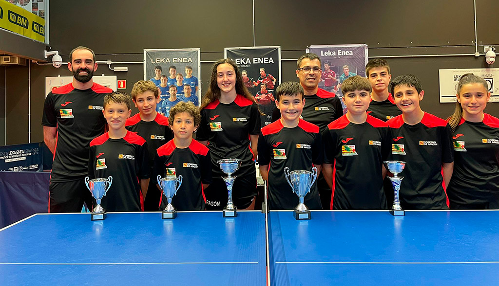 Varios niños y niñas y sus entrenadores posan delante de una mesa de tenis de mesa en representación de la selección de aragón, en la copa pirineos