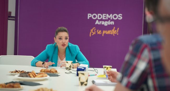 Maru Díaz de Podemos Aragón, sentada en una mesa con el logo de podemos al fondo, con un desayuno al que ha invitado a los periodistas