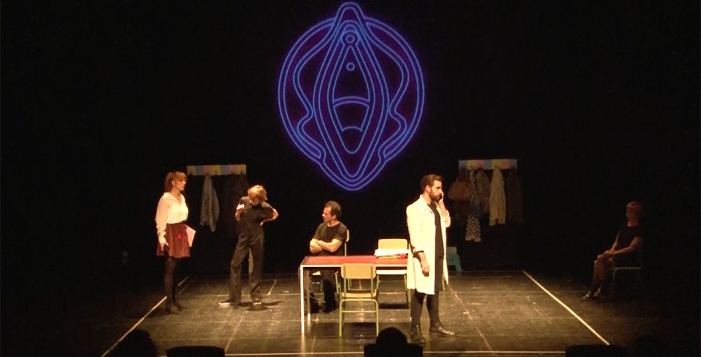 Escenografía de Vulva con cuatro actores y actrices sobre el escenario con una vulva gigante de neón azul