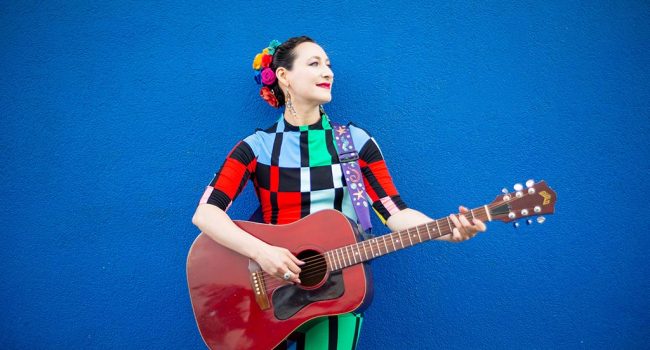 Rachel Sage con una guitarra en un fondo azul