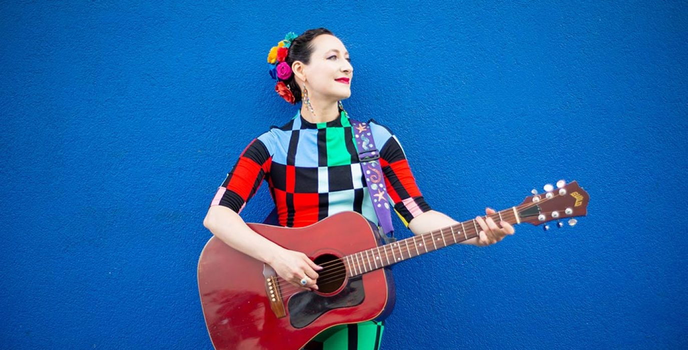 Rachel Sage con una guitarra en un fondo azul
